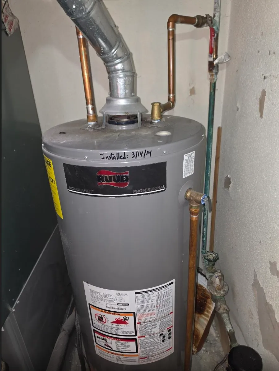 12-year-old Ruud gas water heater marked installed 3/14/14, with a green, corroded supply pipe and valve beside it